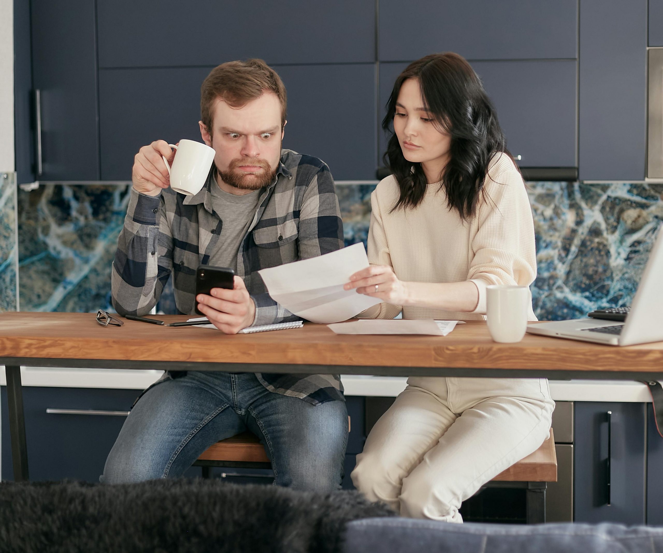 man with a shocked expression holding a coffee mug while looking at the bill his wife is holding
